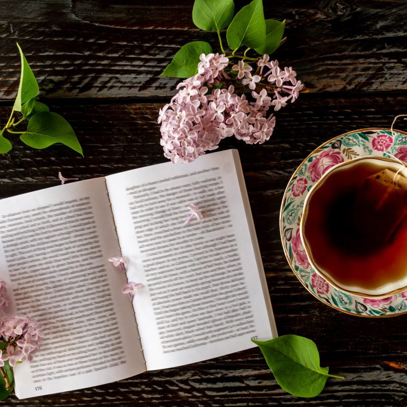 Top view of open book in English and cup of black tea with lilac on wooden table. Romantic summer background Top view of open book in English and cup of black tea with lilac on wooden table. Romantic summer background.